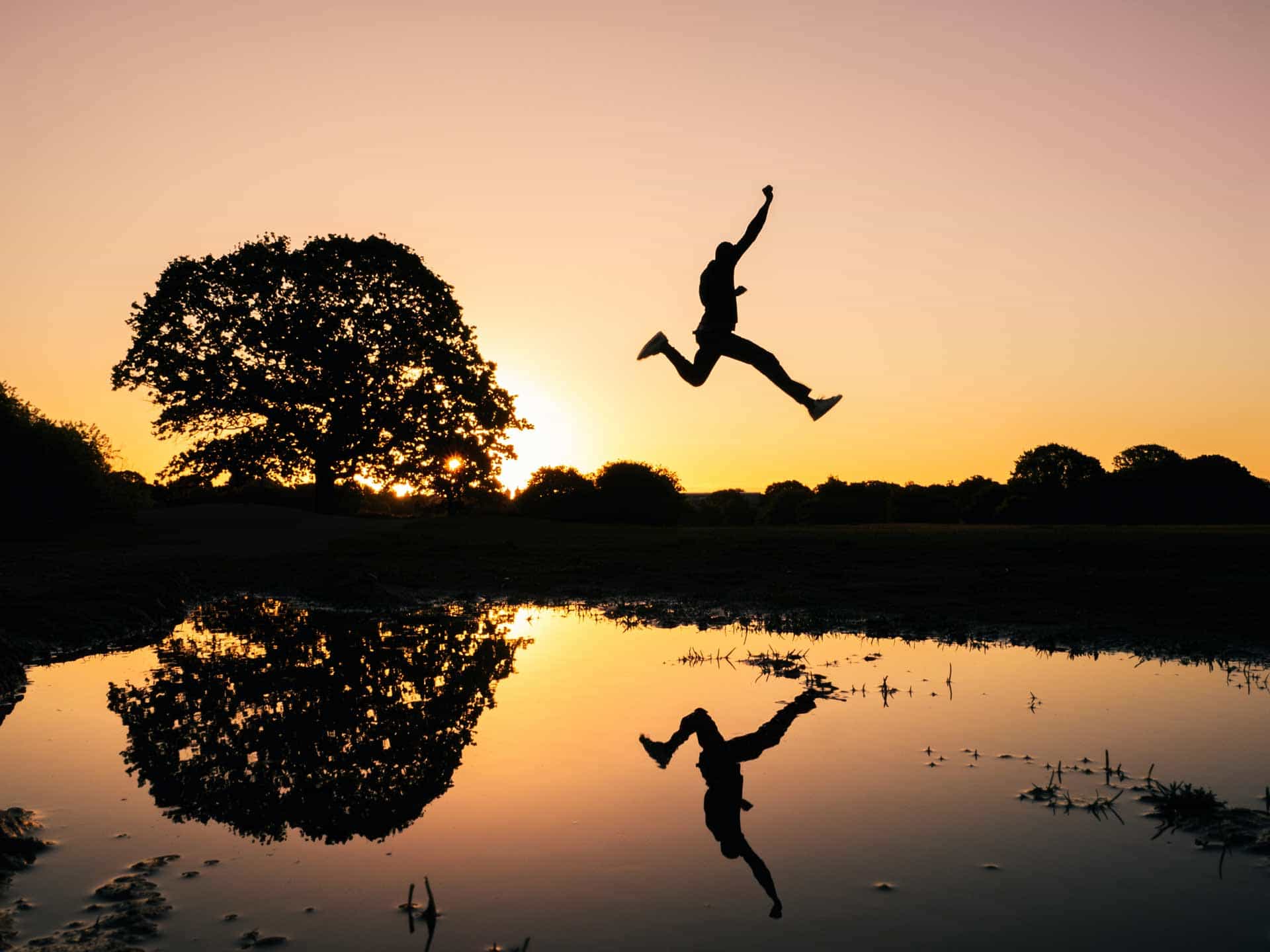 The silhoutte of a man jumping over water, his body perfectly reflected in the pond below.