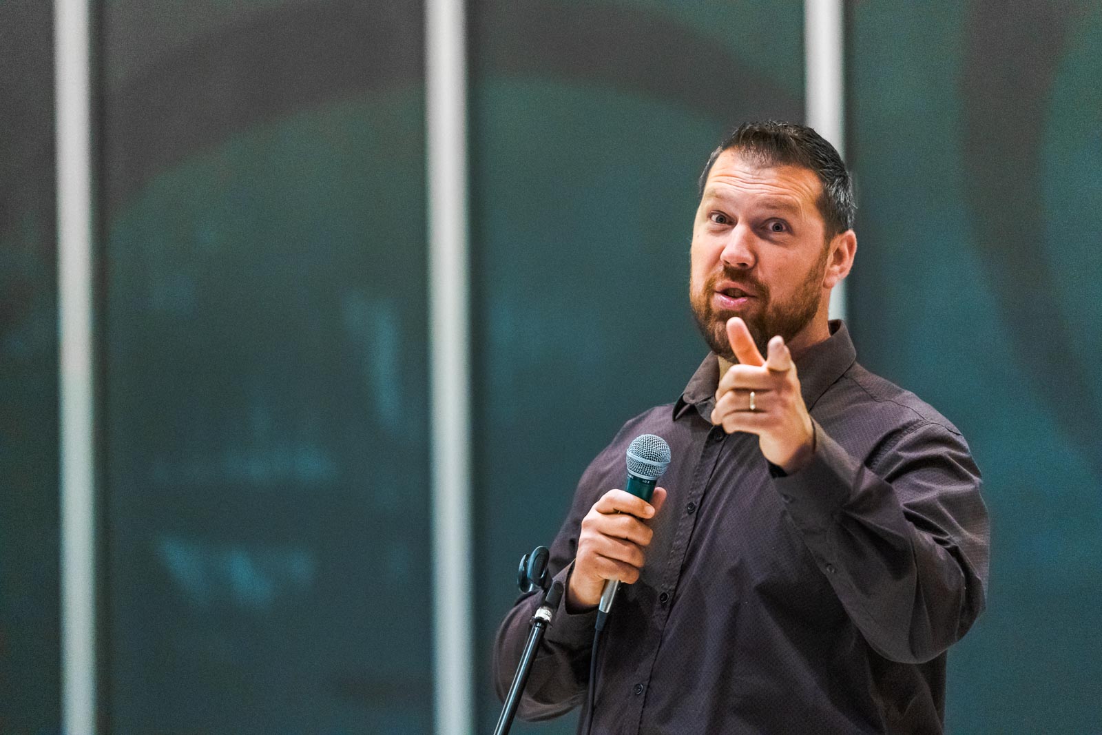 A man stands in front of large windows, pointing his finger at the camera mid-speech.