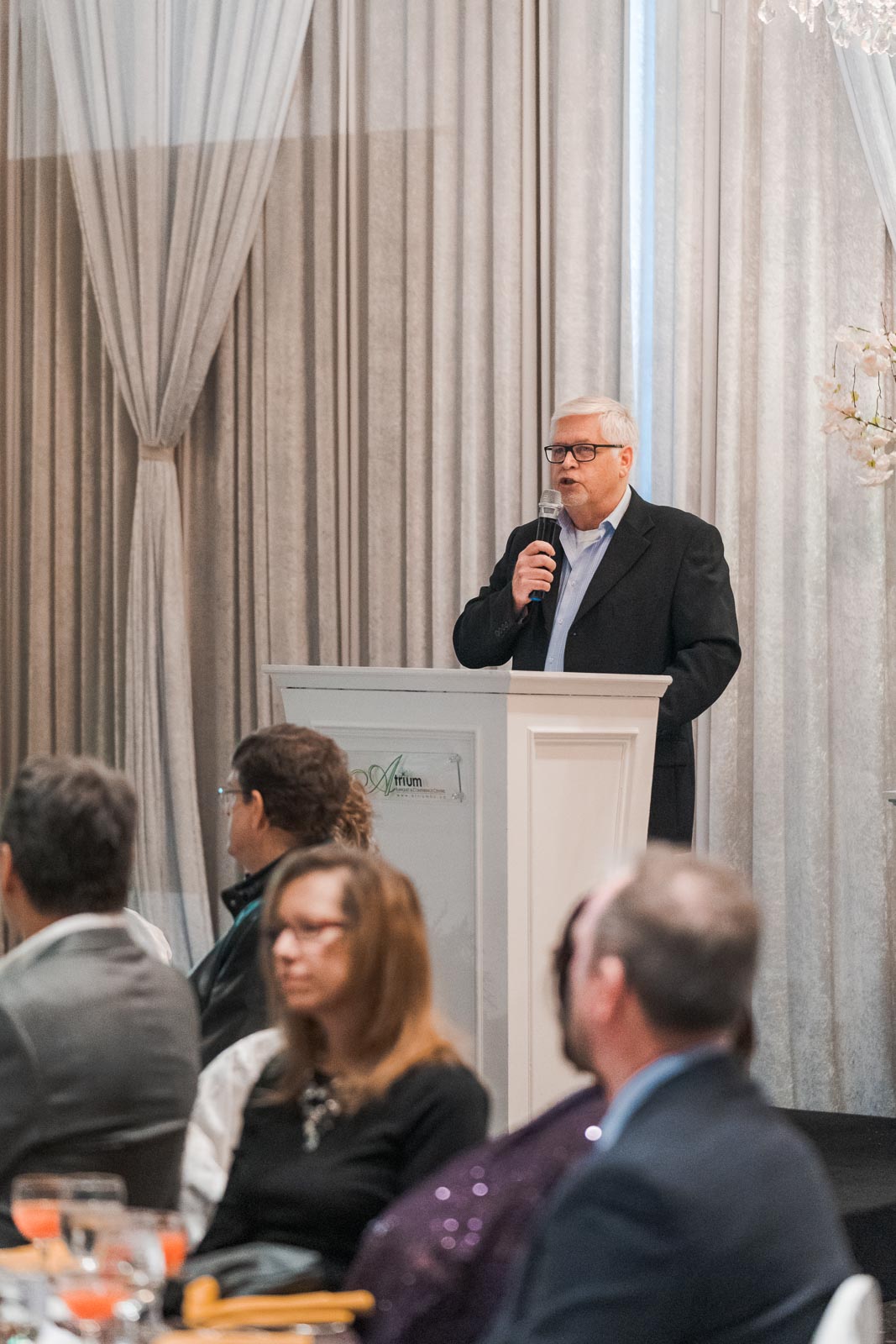 A man stands in front of a podium, holding a mic, speaking mid-sentence to a small group of onlookers seated at tables.