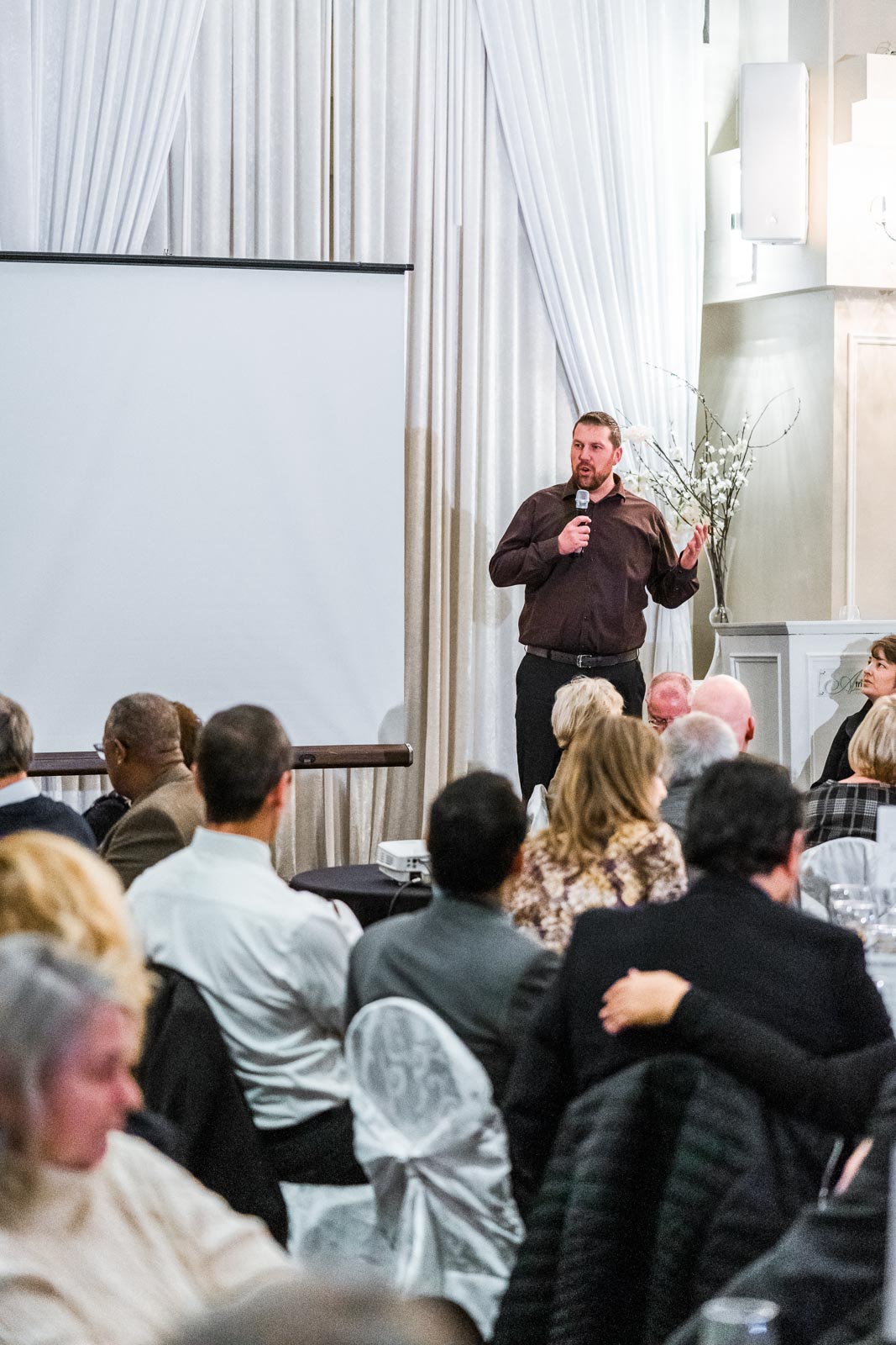 A man energetically speaks on stage, standing beside a projector screen,