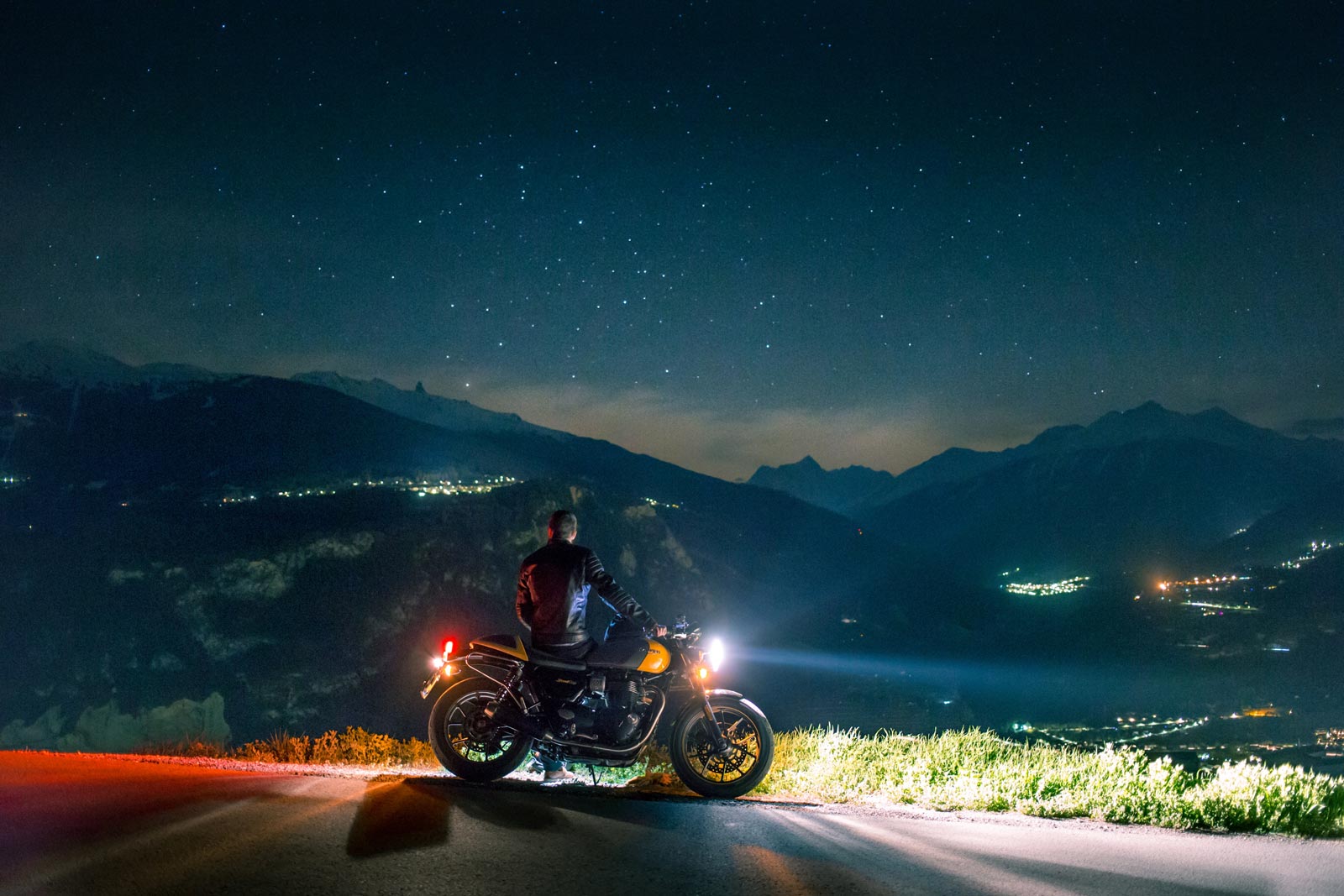 A man sits on a motorcycle and stares out at a star-filled night sky, mountains in the distance.